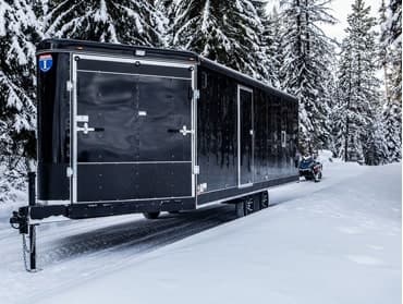 A TrailersPlus trailer in a snowy setting with tall evergreen trees in the background.